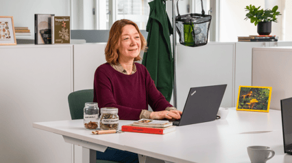 En dame sitter i et kontorlandskap foran en PC og ser tankefullt ut i luften. Rundt henne ligger soppbøker og tørket sopp