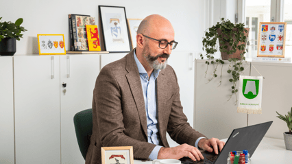 En mann med skjegg sitter i et kontorlandskap foran en PC.