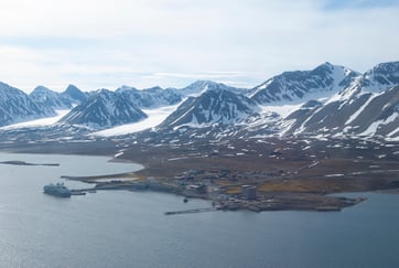 Kystsamfunn i arktis omgitt av snødekte fjell, med en cruiseskip ved kai, spredte bygninger langs strandlinjen, og rolig vann under delvis skyet himmel.