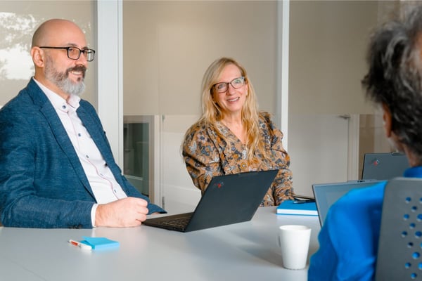 Tre personer sitter rundt et møterombord med bærbare PC‑er foran seg. Det ligger notatlapper, en kopp og en bok på bordet.
