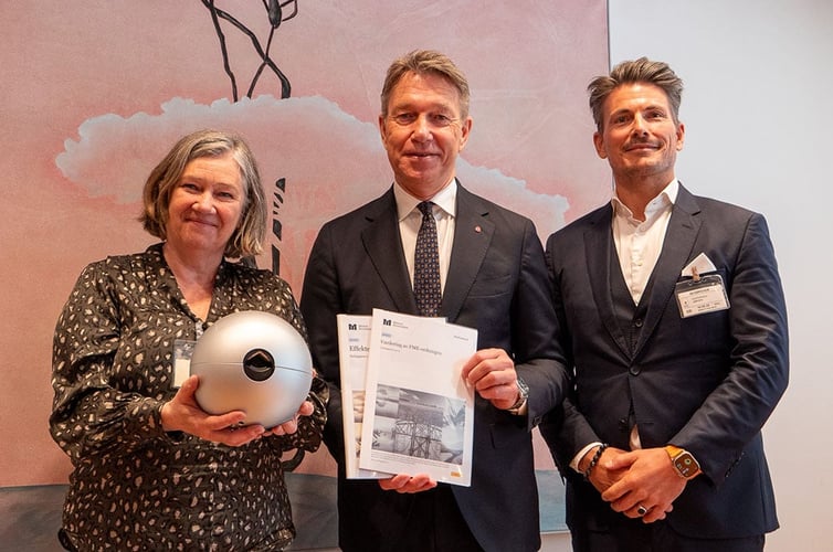 Tre personer poserer foran et maleri. Personen til venstre holder en rund, sølvfarget gjenstand, mens personen i midten holder to rapporter merket med logoen til Norges forskningsråd. Alle tre er pent kledd og smiler mot kameraet, trolig i forbindelse med en offisiell overrekkelse eller presentasjon.