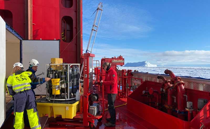 tre mennesker ombord på et forskningsfartøy og noen måleinstrumenter, blå himmel og isfjell i bakgrunnen
