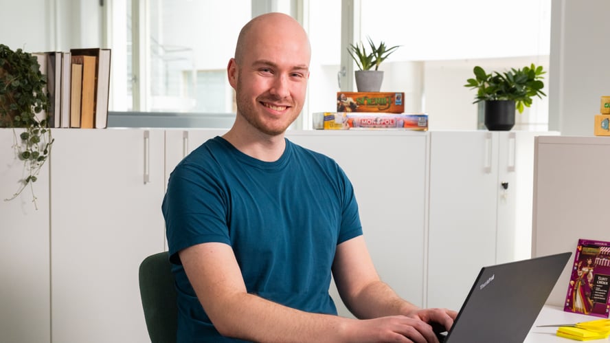 En ung mann sitter foran en PC i et kontorlandskap. Han smiler mot kamera