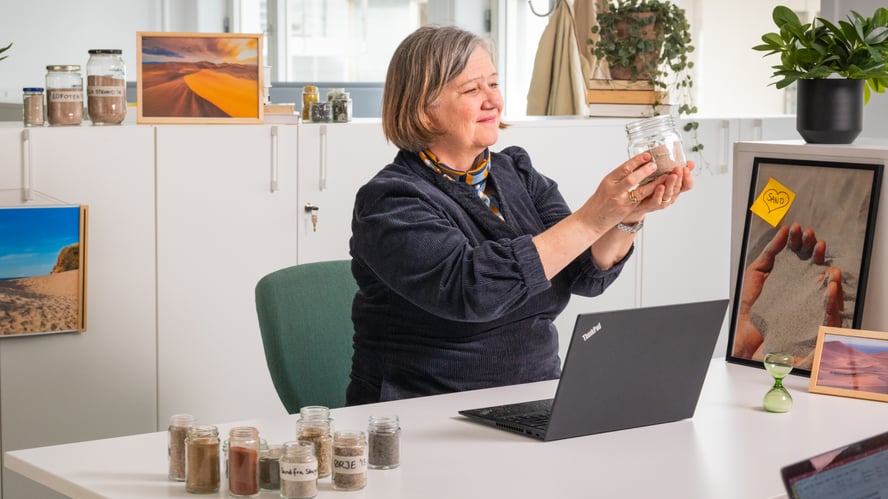En smilende dame sitter i et kontorlandskap foran en PC og holder opp et norgesglass med sand